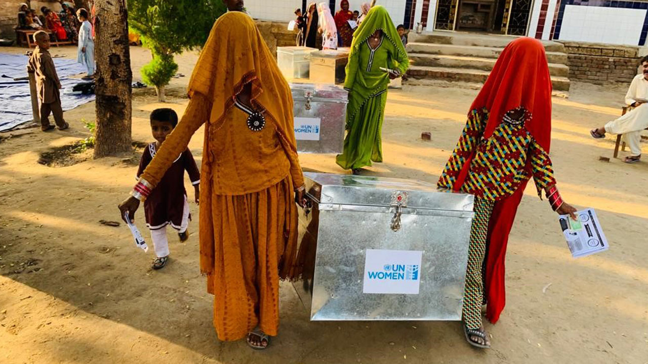 Two Pakistanii women carrying a metal box with UN Women logo is shown clearly.