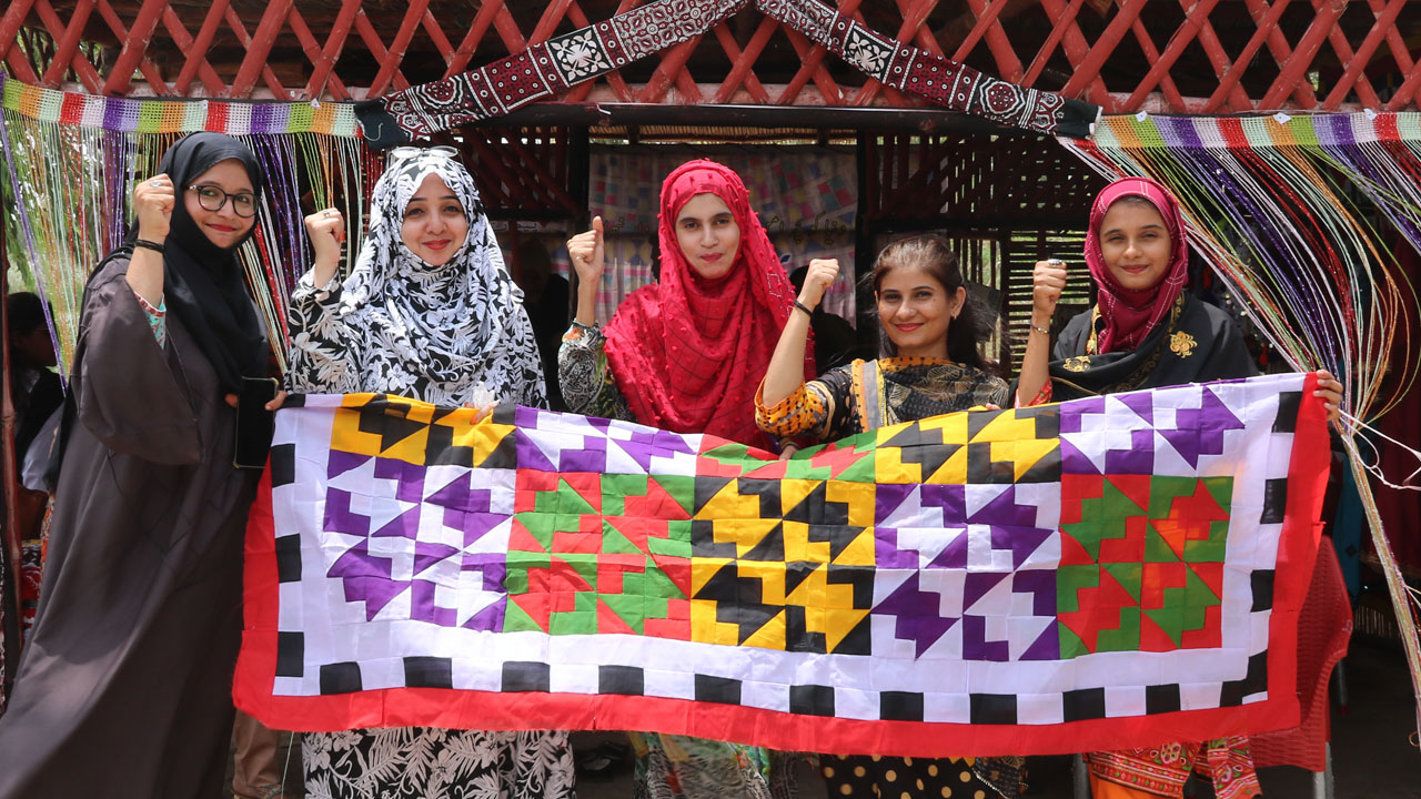 Five Pakistanii women standing proud pulling a hand-made carpet that they just finished making.