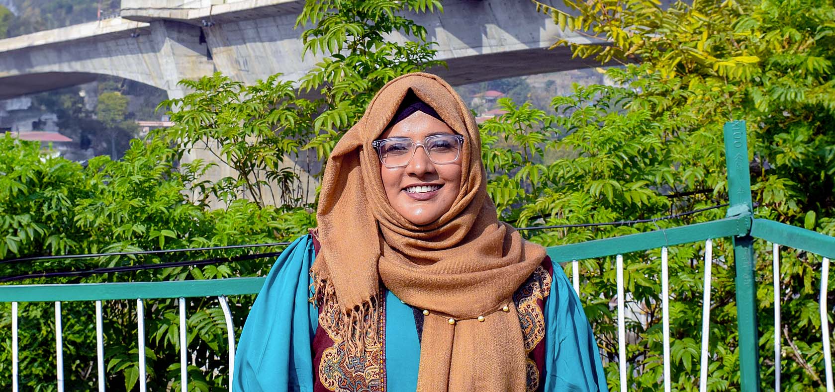 A smiling woman with glasses and a hijab, wearing a teal dress with a brown scarf, sits outside with greenery and a bridge in the background.  Photo courtesy of Maryam Eqan
