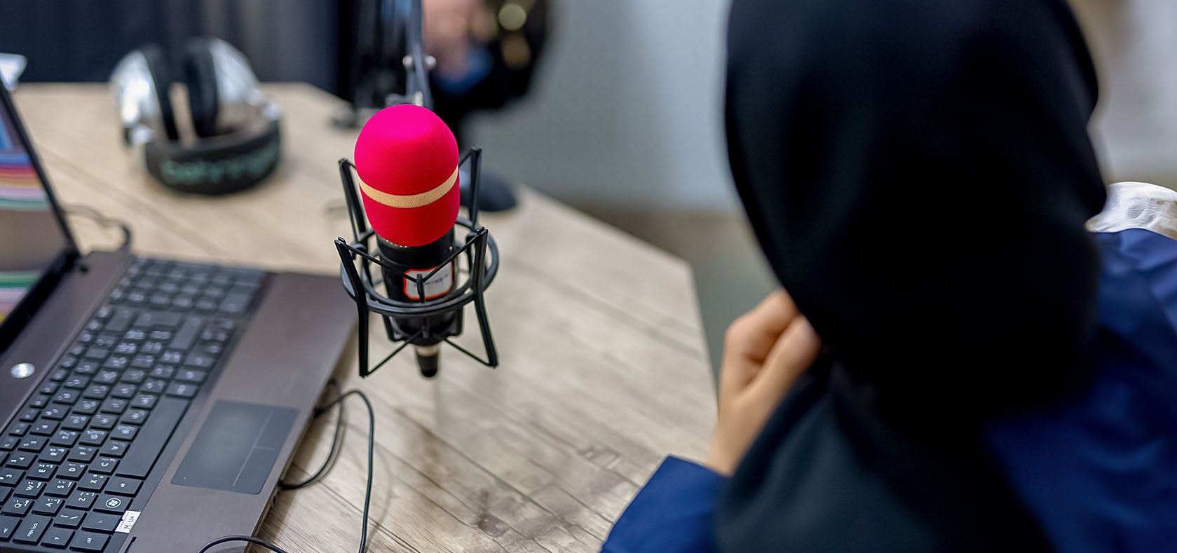A Muslim woman in a black burqa is seen from her backside sitting while talking over a red microphone that is connected to a laptop PC.