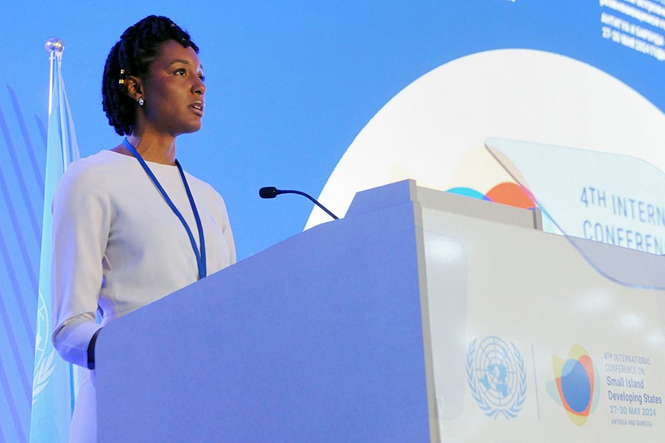 A young dark-skinned woman from an island wearing a white shirt standing at the podium dsdelivering her lecture to the cloud.
