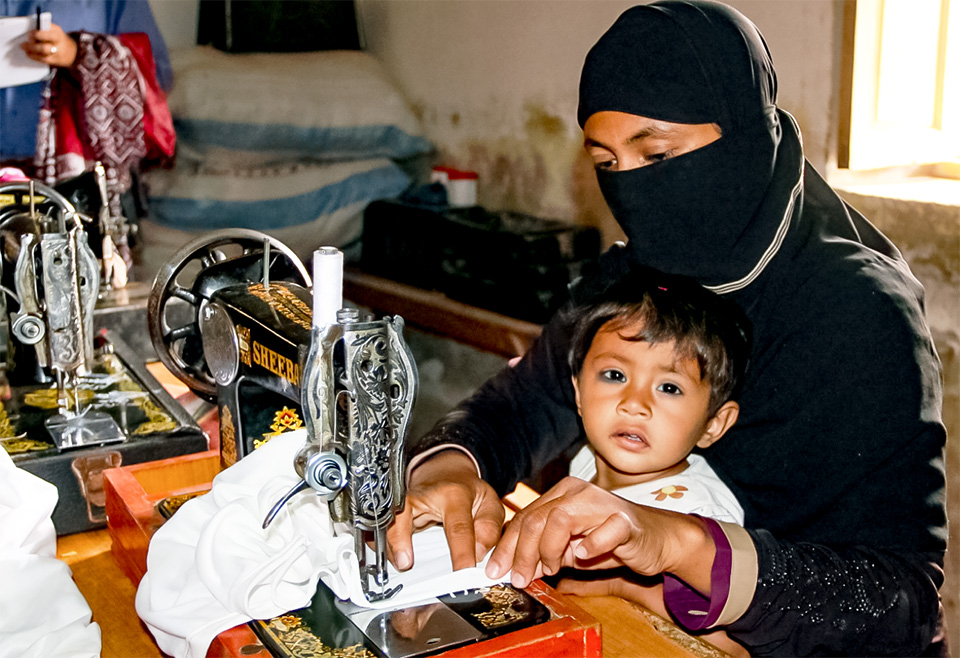 Salma works on sewing machine at Community Center in Gharo, while her son looks on. Photo: UN Women/Habib Asgher