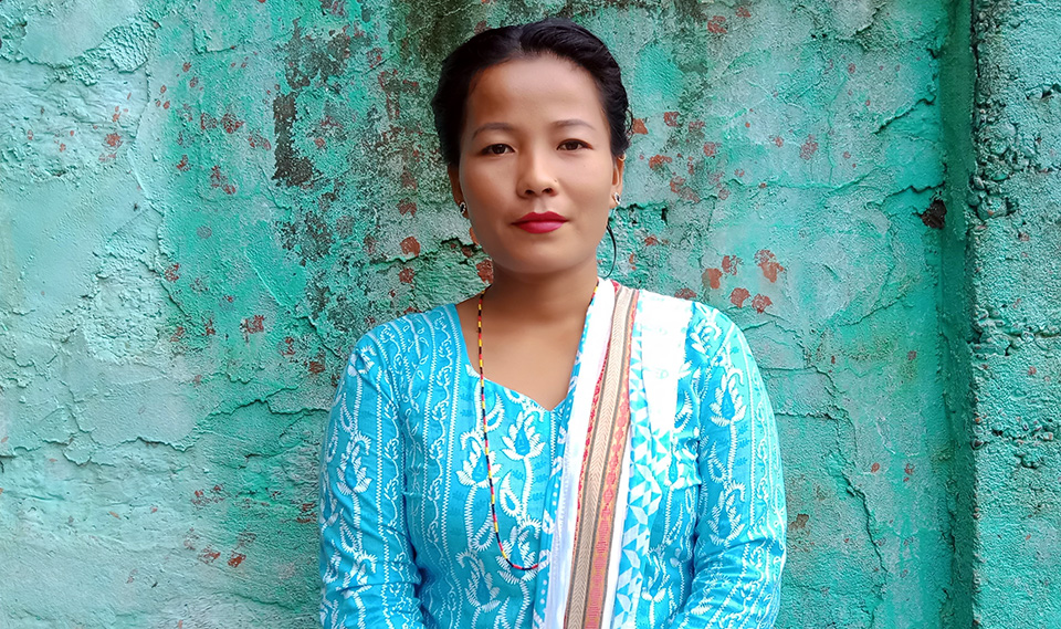 Photo of Shila, waring blue Nepali shirt standing against cement wall also painted in blue. Photo: Courtesy of Shila Ale