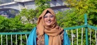 A smiling woman wearing glasses and a hijab, in a teal dress with a brown scarf, sitting outdoors with greenery and a bridge in the background. Photo courtesy of Maryam Eqan